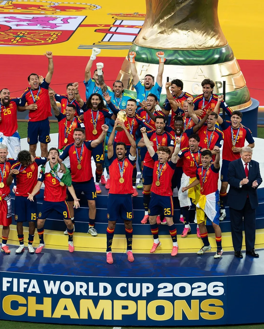Spain players celebrate on the podium with the World Cup trophy under a "FIFA World Cup 2026 Champions" sign